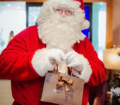 Santa Claus delivering gifts in festive hotel lobby with twinkling lights and Christmas tree, spreading holiday cheer.