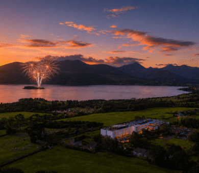 Stunning sunset view over lake and hills with fireworks, welcoming hotel, lush grounds, perfect for a serene getaway.