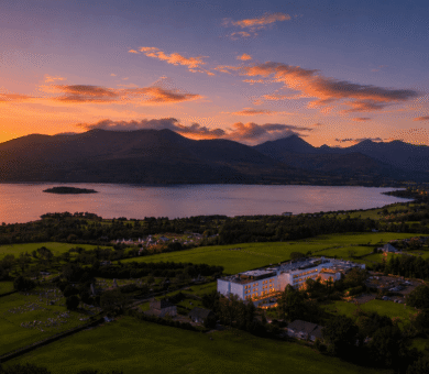 Stunning sunset view over lake and mountains with illuminated hotel amidst lush greenery, perfect for a serene getaway.