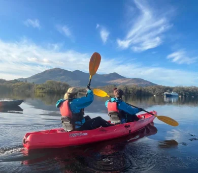 Guests kayaking on tranquil lake with scenic mountain views and clear blue skies; adventure activities at countryside hotel.